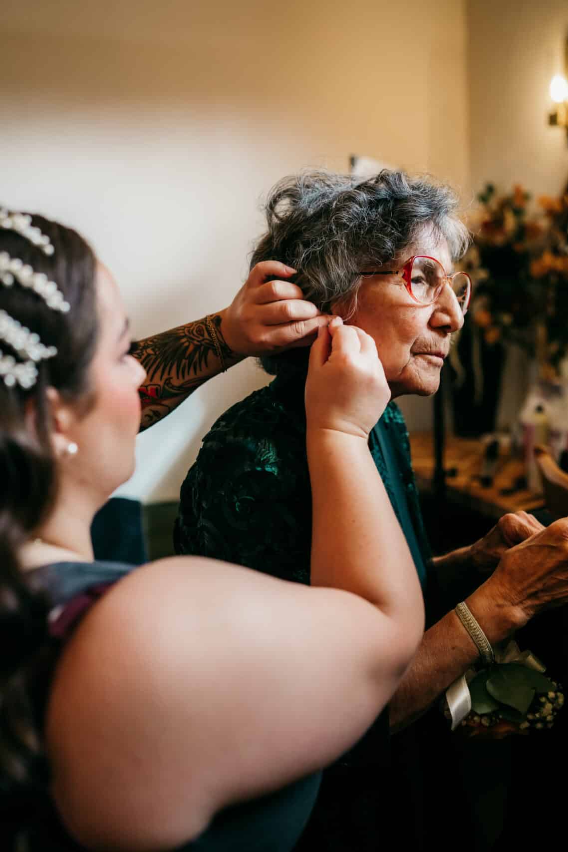 Elegant woman helping elderly woman with earrings at special event, capturing intimate family moment.