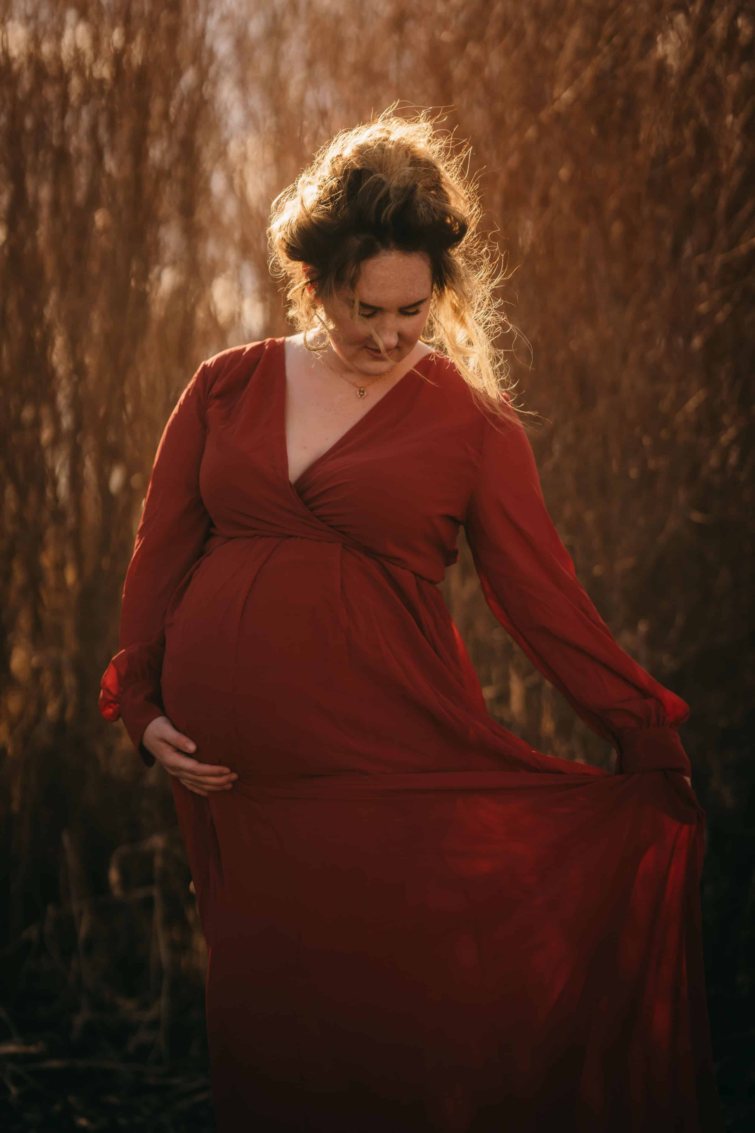Pregnant woman in a flowing red dress outdoors during golden hour, embracing her pregnancy and showcasing maternal beauty, surrounded by warm natural lighting.