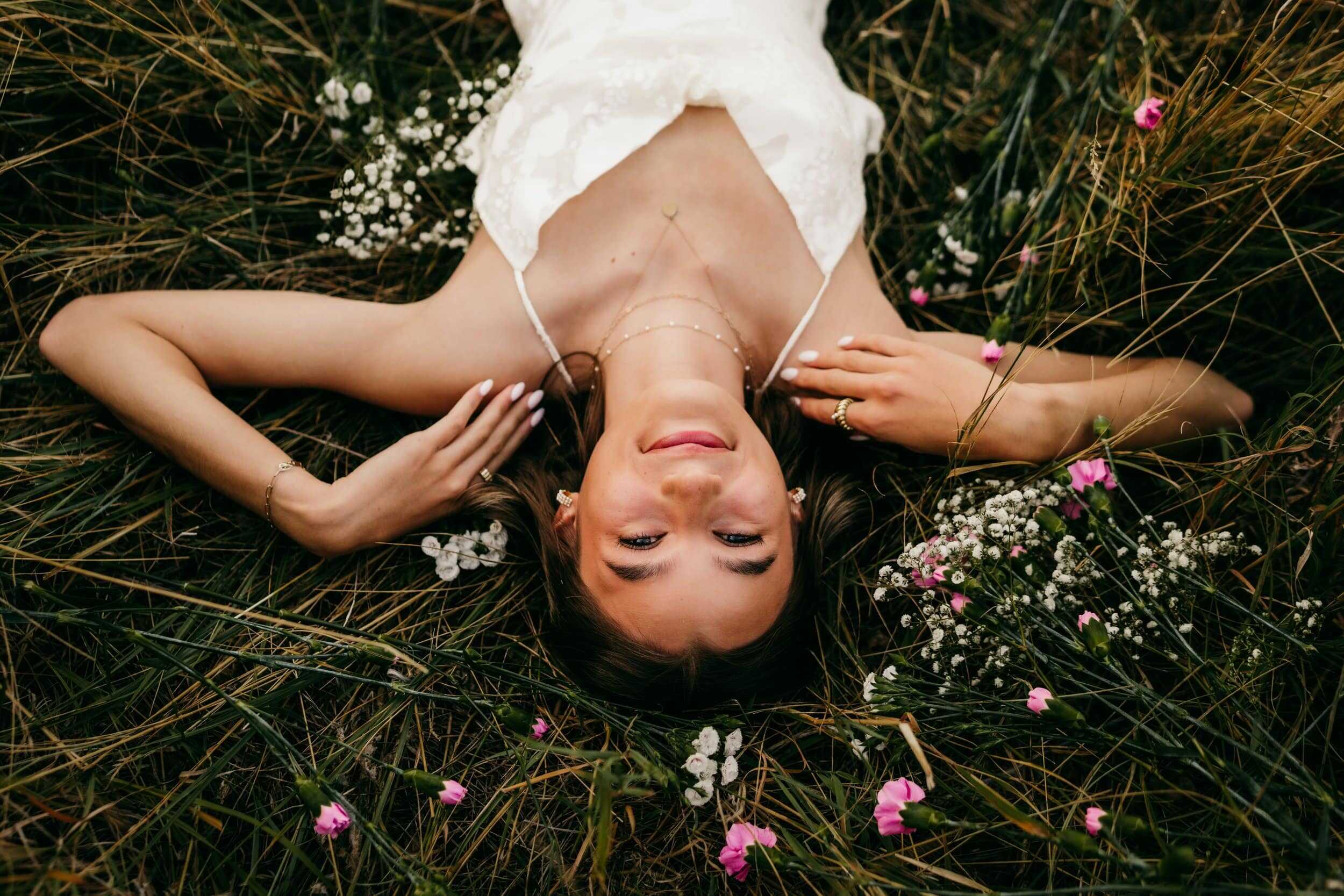 Elegant woman lying on grass with wildflowers, wearing a white dress and jewelry, captured in a serene, outdoor portrait emphasizing natural beauty and fashion.