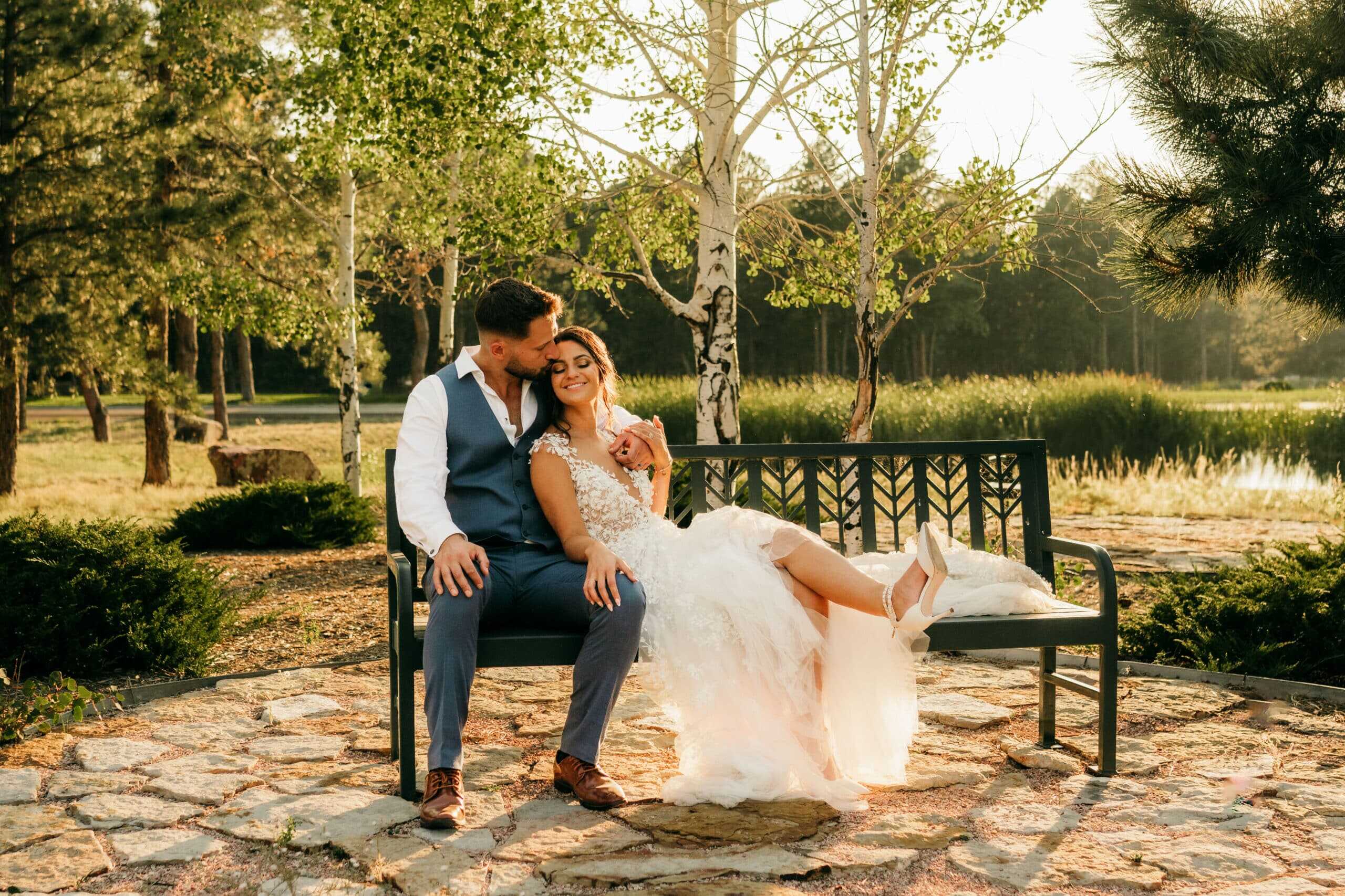 Romantic wedding couple sitting on bench in a scenic park during sunset, with lush greenery and a pond in the background.