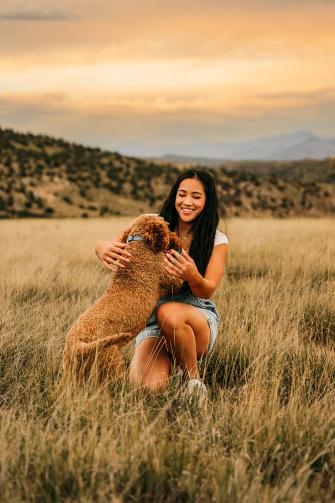 Cute woman playing with a curly brown poodle dog outdoors in a grassy field at sunset, scenic mountain background, joyful moment, pet companionship, outdoor photography.