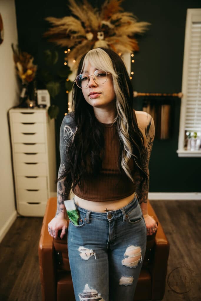 Vibrant woman with black and blonde hair, tattoos, glasses, wearing a brown crop top and ripped jeans, posing indoors with decorative warm lighting and foliage backdrop.