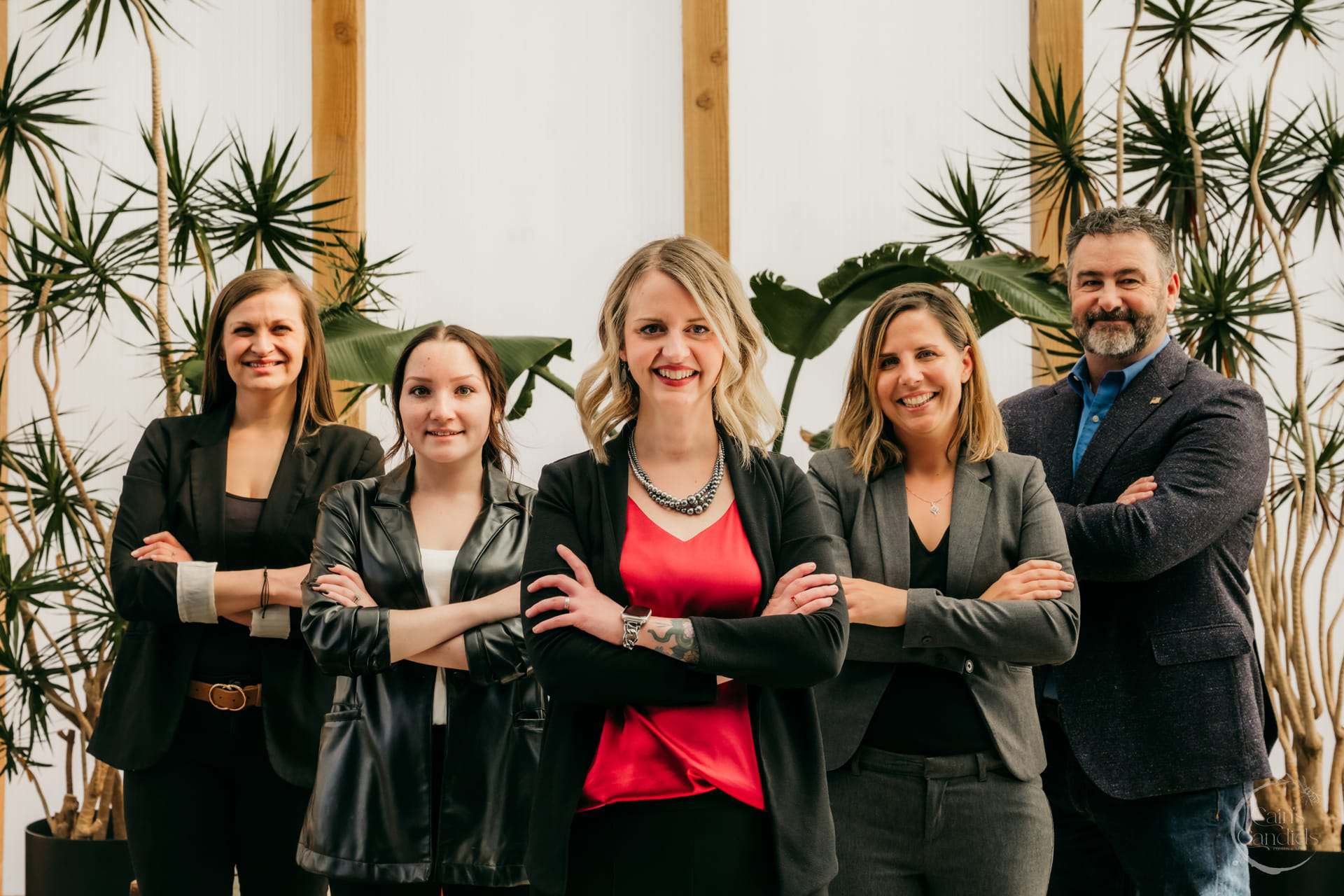 Confident professional team portrait featuring five diverse colleagues standing together with arms crossed, inside a modern office with lush indoor plants, showcasing teamwork, leadership, and success.