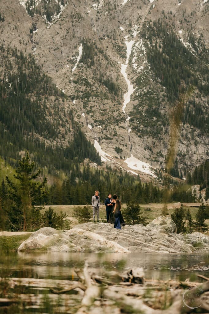 Elegant wedding ceremony outdoors in front of majestic mountain landscape, with couples exchanging vows on rocky outcrop near serene water, surrounded by lush forest and scenic natural beauty.